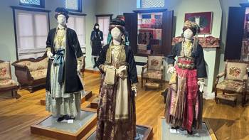 Mannequins dressed with traditional Greek women's costumes in a museum room with vintage furniture and embroidered chairs in the background.